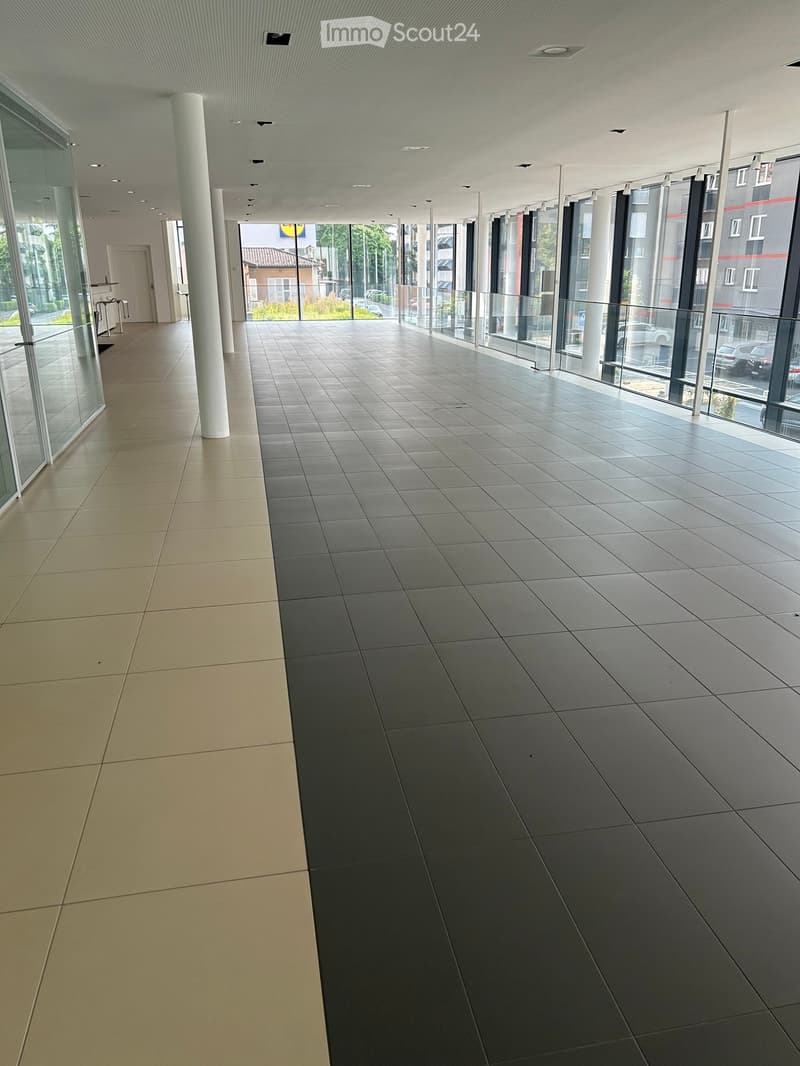 Modern hallway with tiled floors, large glass windows, and white columns. The space is well-lit with recessed ceiling lights and features a clean, minimalist design.