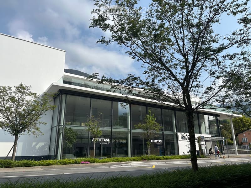 Modern two-story building with large glass windows and a white facade, surrounded by trees and greenery.