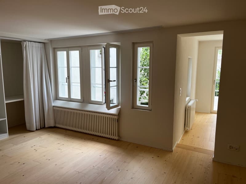 Empty room with light wooden flooring, large windows with white curtains, radiators, and a doorway leading to another space.