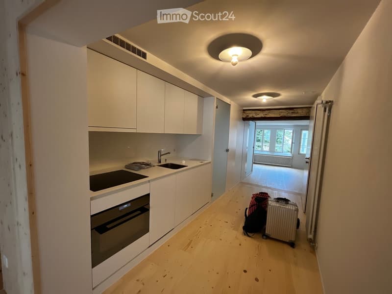 A narrow hallway with light wooden flooring, white cabinets, and a modern kitchen area visible on the left. A suitcase and backpack are placed on the floor near the doorway leading to another room.
