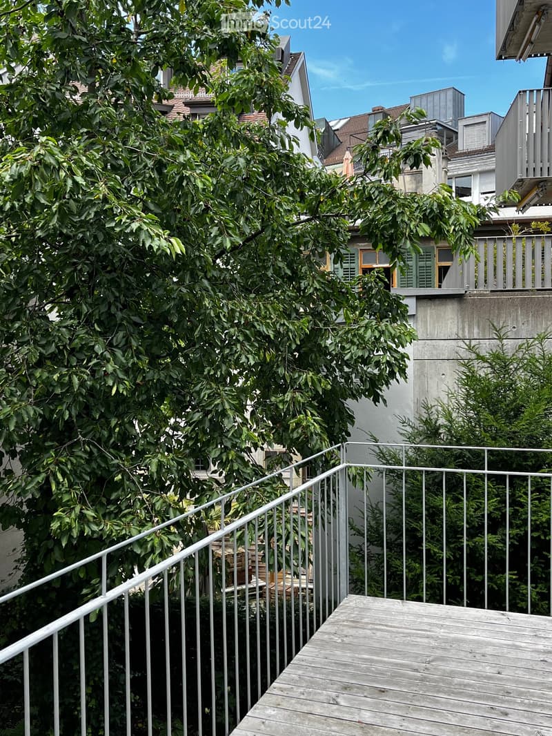 Wooden balcony with metal railing, surrounded by lush green trees and neighboring buildings.