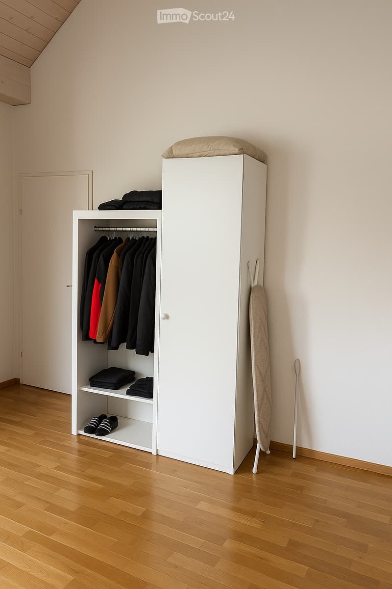 A minimalist, unfurnished room with a white wardrobe, clothes hanging inside, a shelf with folded blankets, and shoes on the bottom shelf. The room features light wooden flooring and white walls.