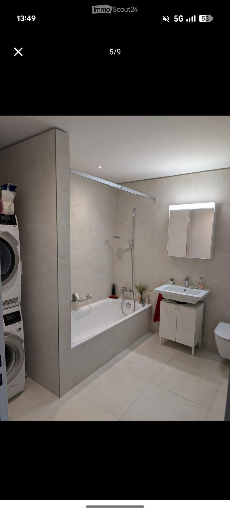 Modern bathroom with a bathtub, sink, mirror, toilet, and a washing machine in the corner. Features a sleek design with beige tiles and a built-in showerhead.