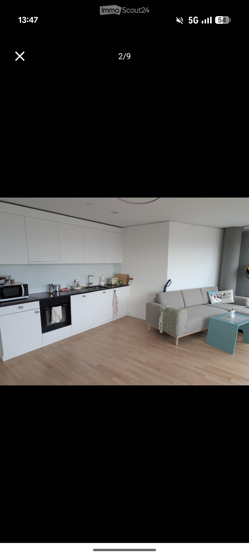 Modern open-plan living and kitchen area with light-colored cabinets, a grey sectional sofa, a small blue coffee table, and a modern kitchen setup including a microwave and oven.