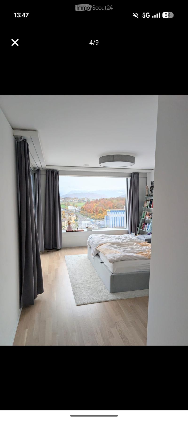 Modern bedroom with wooden flooring, large window offering scenic view, bed with white and orange bedding, shelf with books, and decorative items on windowsill.
