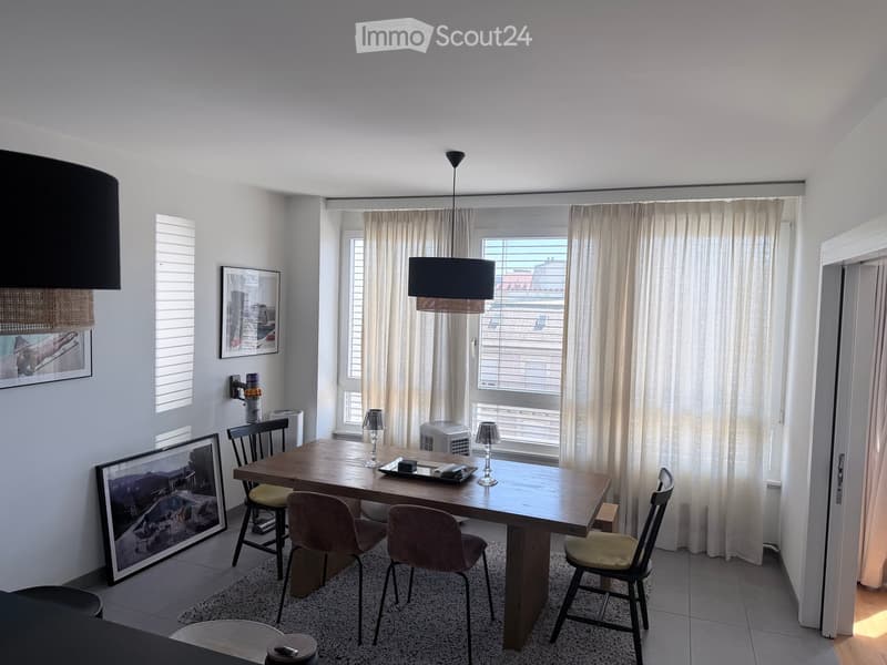 Bright dining area with a wooden table, four chairs, and a hanging lamp. Large windows with curtains and framed artwork on the walls.