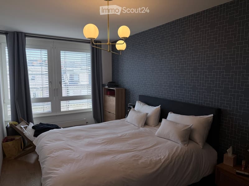 Modern bedroom with a large bed, nightstands, a wooden chair, and a patterned wall design. The room features large windows with blinds and a stylish chandelier.