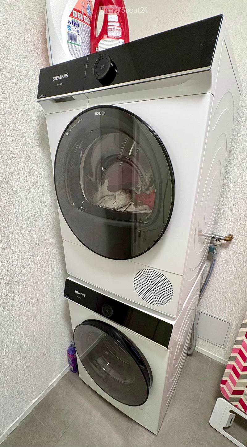 A modern bathroom with a white Siemens washing machine and a dishwasher stacked vertically, with cleaning supplies visible on top.