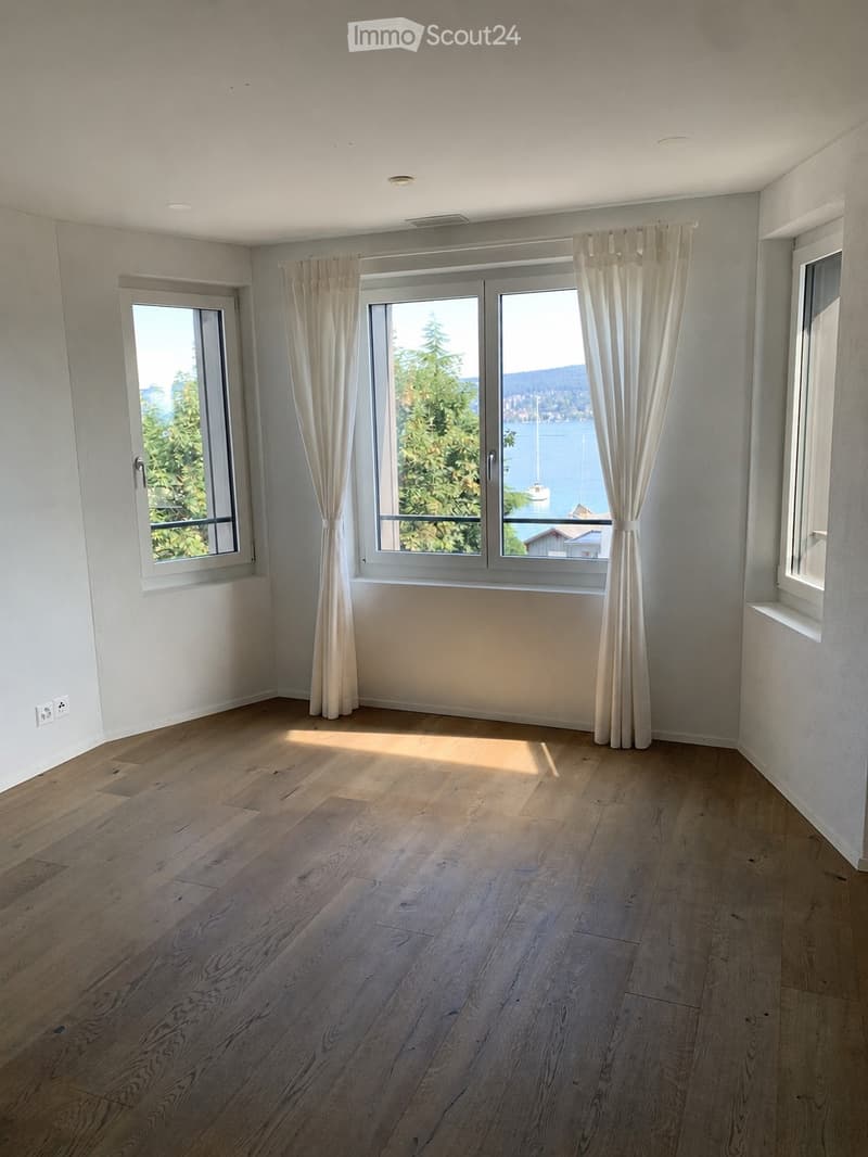 Empty room with light wooden flooring, white walls, large windows with white curtains, and a view of a lake and trees.