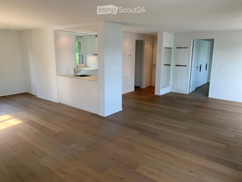 Empty room with light wooden flooring, white walls, and a white kitchen area visible in the background. No furniture or equipment present.