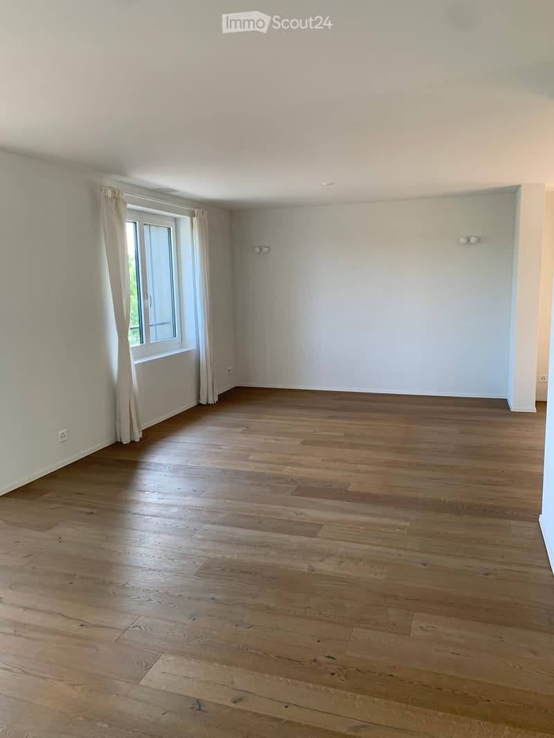 Empty room with light wooden flooring, white walls, and a large window with curtains.