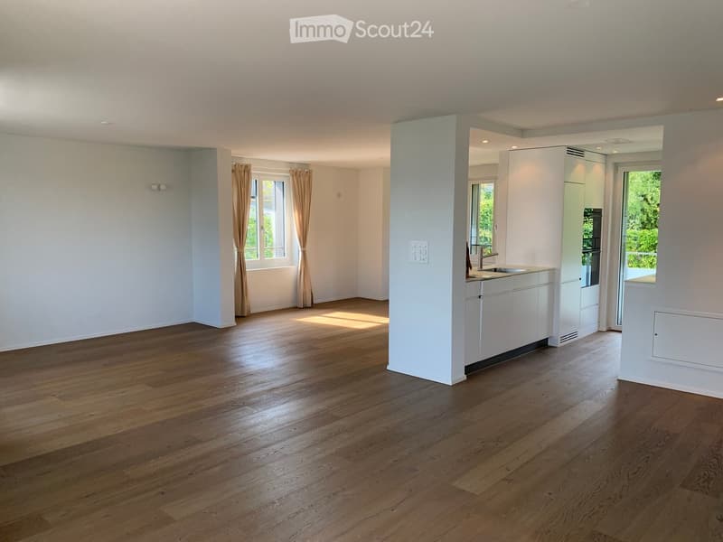 Open-plan living and kitchen area with wooden flooring, white cabinetry, and large windows with curtains.