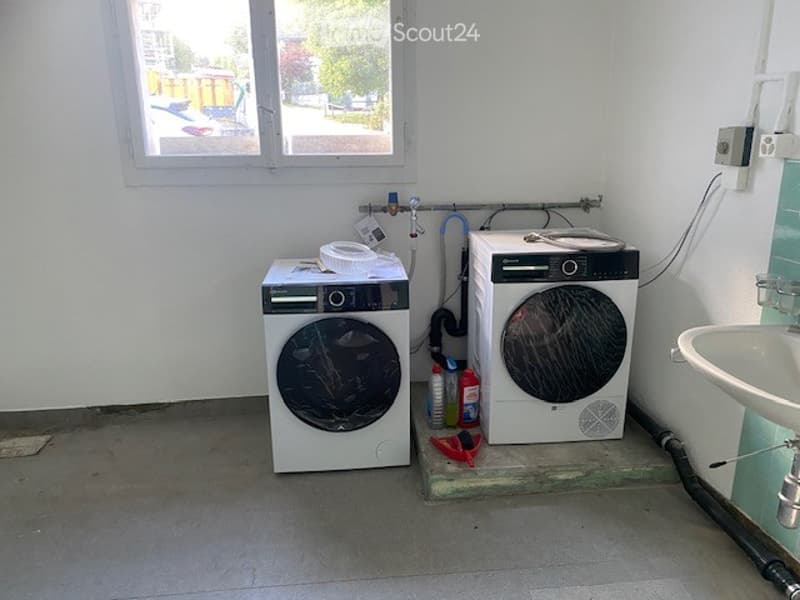 A small utility room with a washing machine, dishwasher, and sink, visible pipes and cleaning supplies.