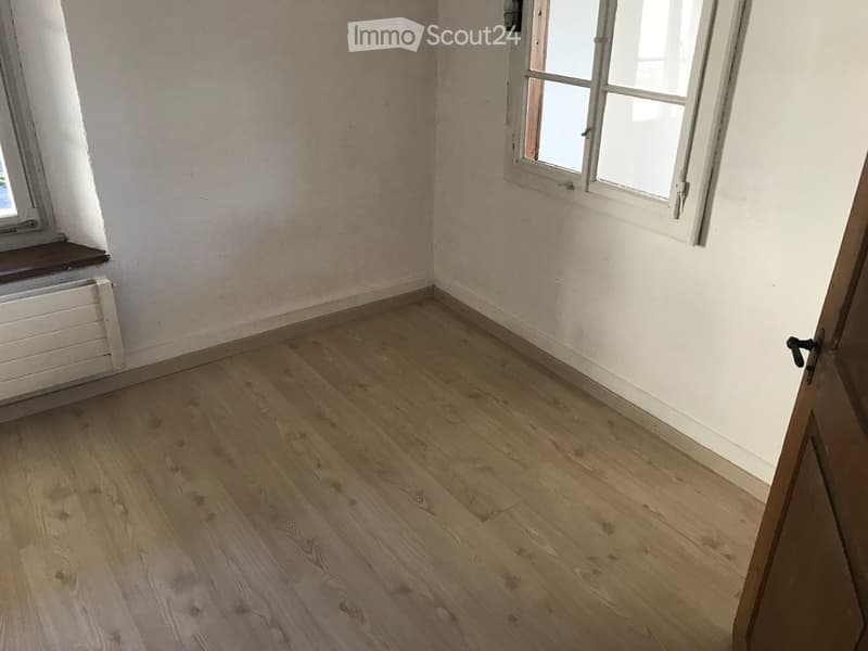 Empty room with wooden flooring, white walls, and two windows with white frames. A radiator is visible under one window, and a door is partially open on the right side.