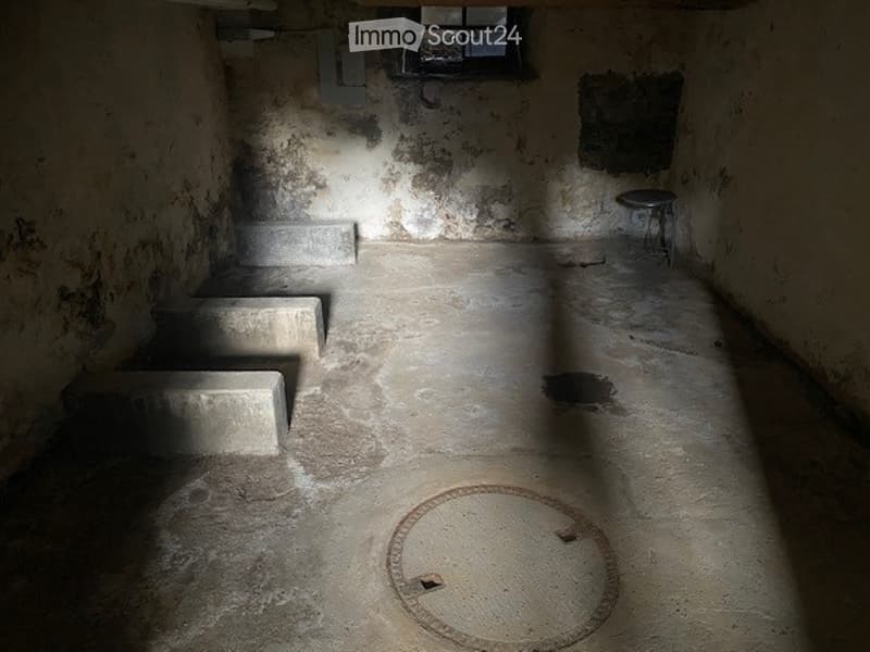 Empty, dilapidated room with exposed concrete blocks, a small window, and a single metal stool. The walls are rough and stained, and the floor is concrete with a manhole cover in the center.