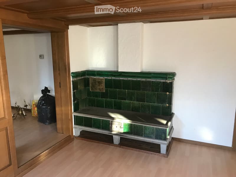 An unfurnished room with a fireplace, wooden floor, and white walls. The fireplace has green tiles and a decorative design.