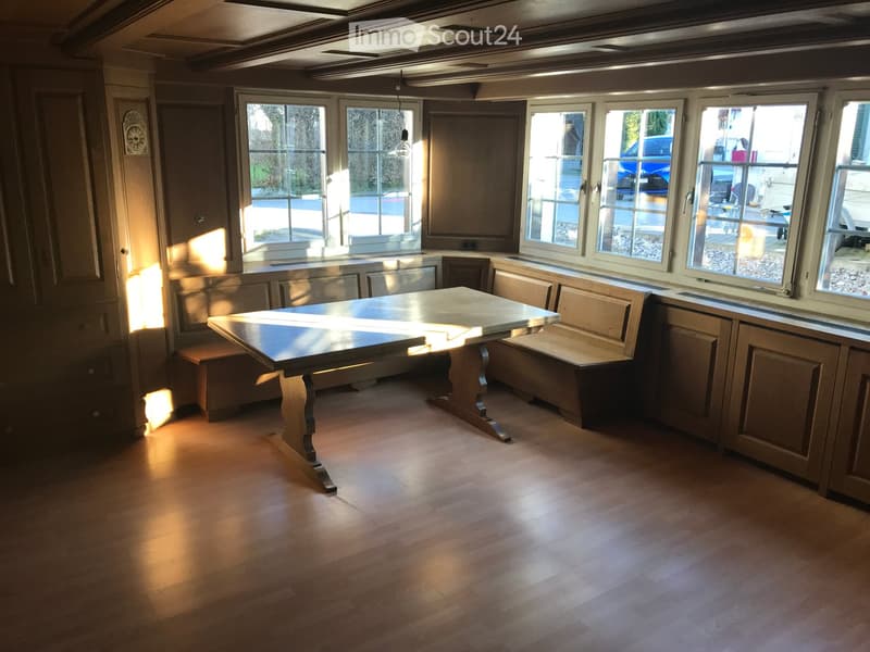 Bright, sunlit room with wooden flooring, a central table, built-in benches, and large windows offering a view of the outside.