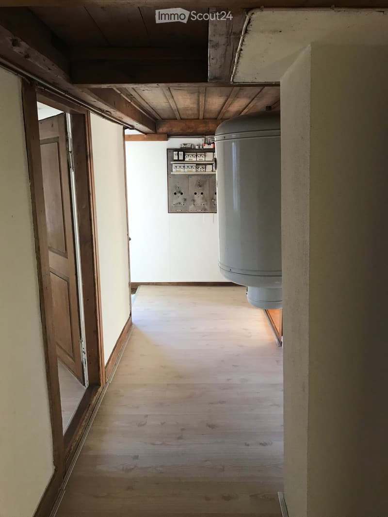 Empty wooden hallway with exposed wooden beams on the ceiling, a grey water tank, and a wooden door on the left side.