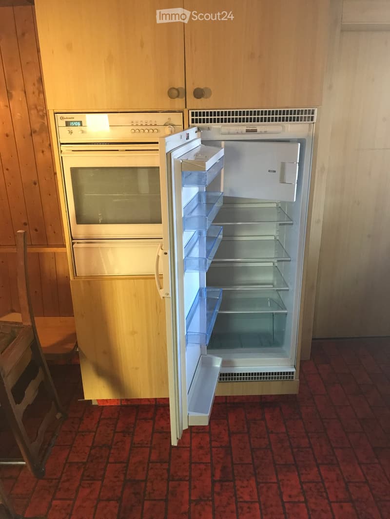A kitchen with a red tiled floor, wooden cabinets, and an open refrigerator next to an oven.