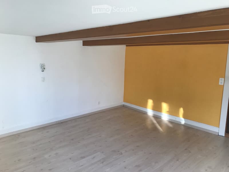 Empty room with light wooden flooring, white walls, and exposed wooden beams on the ceiling. One wall is painted orange.