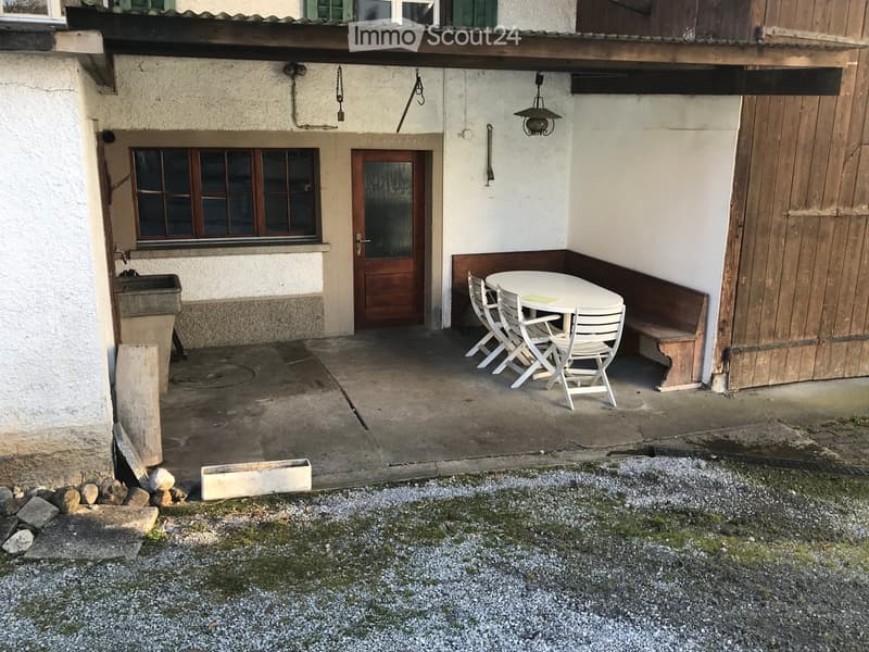 Outdoor seating area with a white table and four white chairs, attached to a house with a wooden door and window.