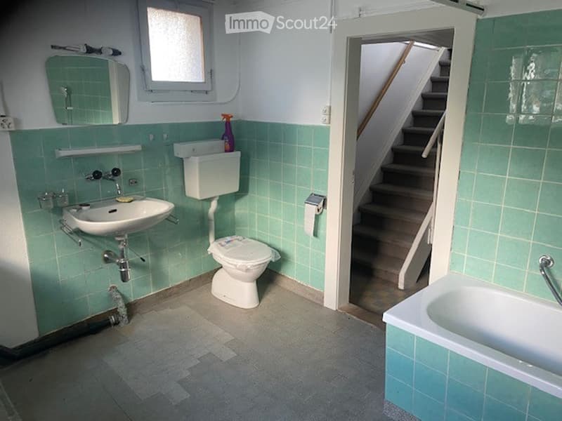 Bathroom with green tiled walls, a sink, toilet, bathtub, and a mirror above the sink. A small window is visible above the sink, and a doorway leads to a staircase.