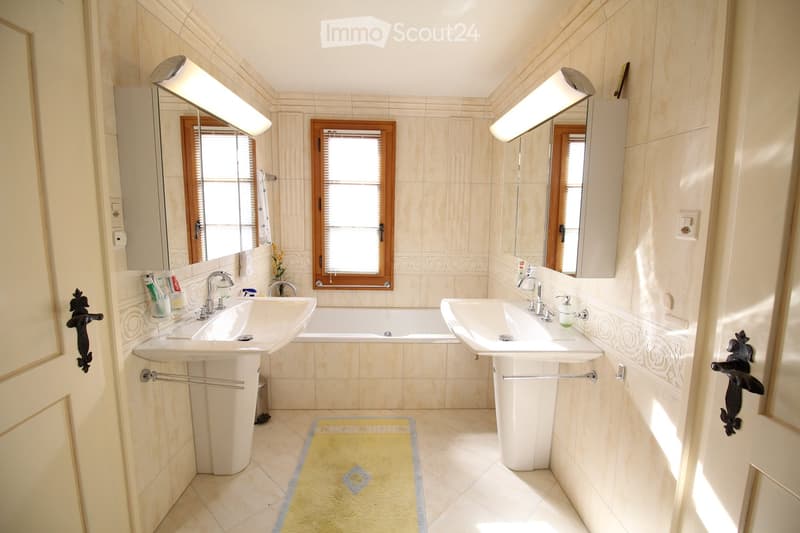 Elegant bathroom with double sinks, a bathtub, large mirrors, and decorative tiles. Features include a window with blinds, towel racks, and a small yellow rug on the floor.