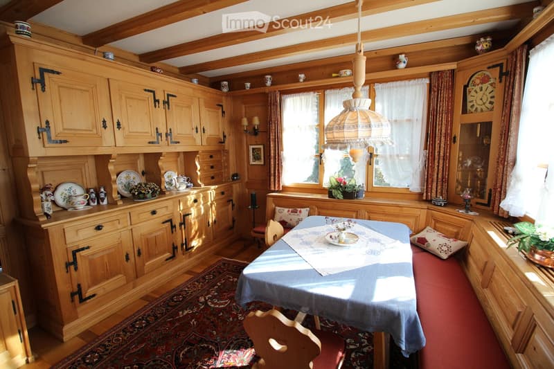 Cozy kitchen with wooden cabinets, a dining table with chairs, decorative plates, teapots, and a flower vase on the windowsill. The room features wooden beams on the ceiling, a patterned rug, and a hanging lamp.