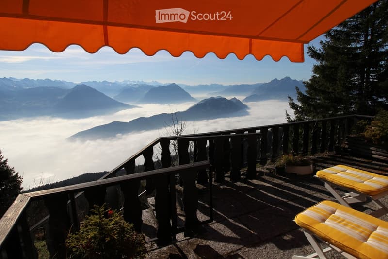 Balcony with a view of mountains and clouds, featuring a yellow cushioned chair and a wooden railing.