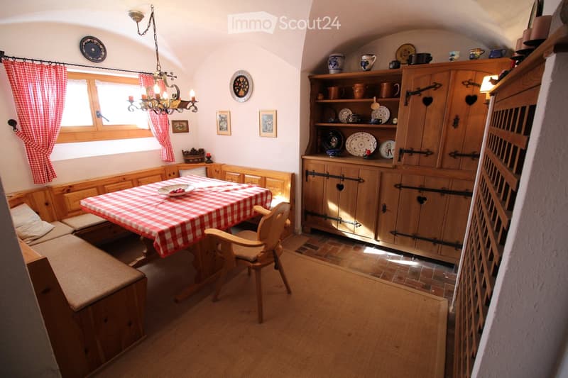 Cozy kitchen with wooden furniture, a dining table with a checkered tablecloth, a wooden chair, and a large wooden cupboard with decorative plates and cups on display.