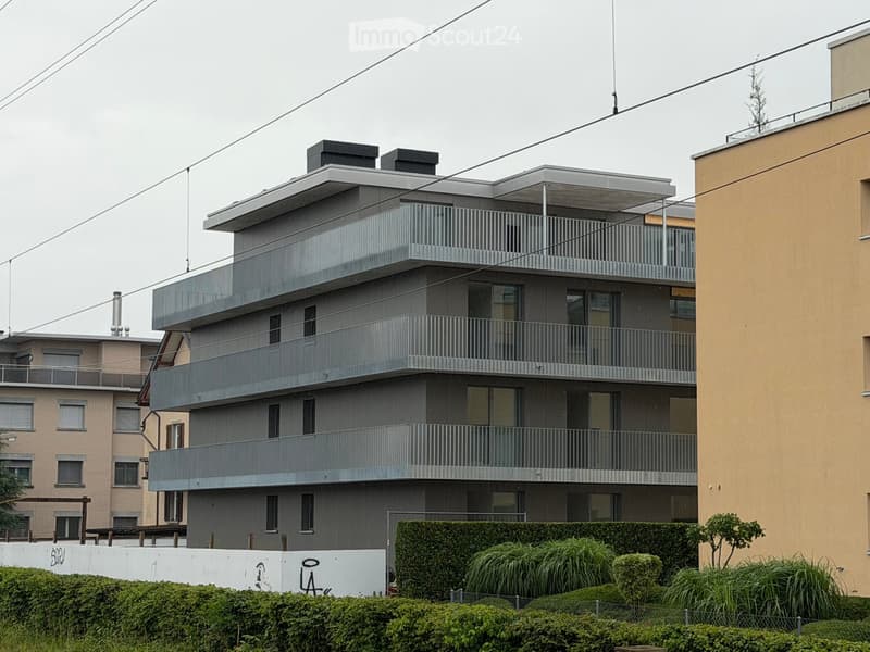 Modern multi-story residential building with grey facade, balconies, and large windows. The building features a sleek, contemporary design with a flat roof and visible chimneys.