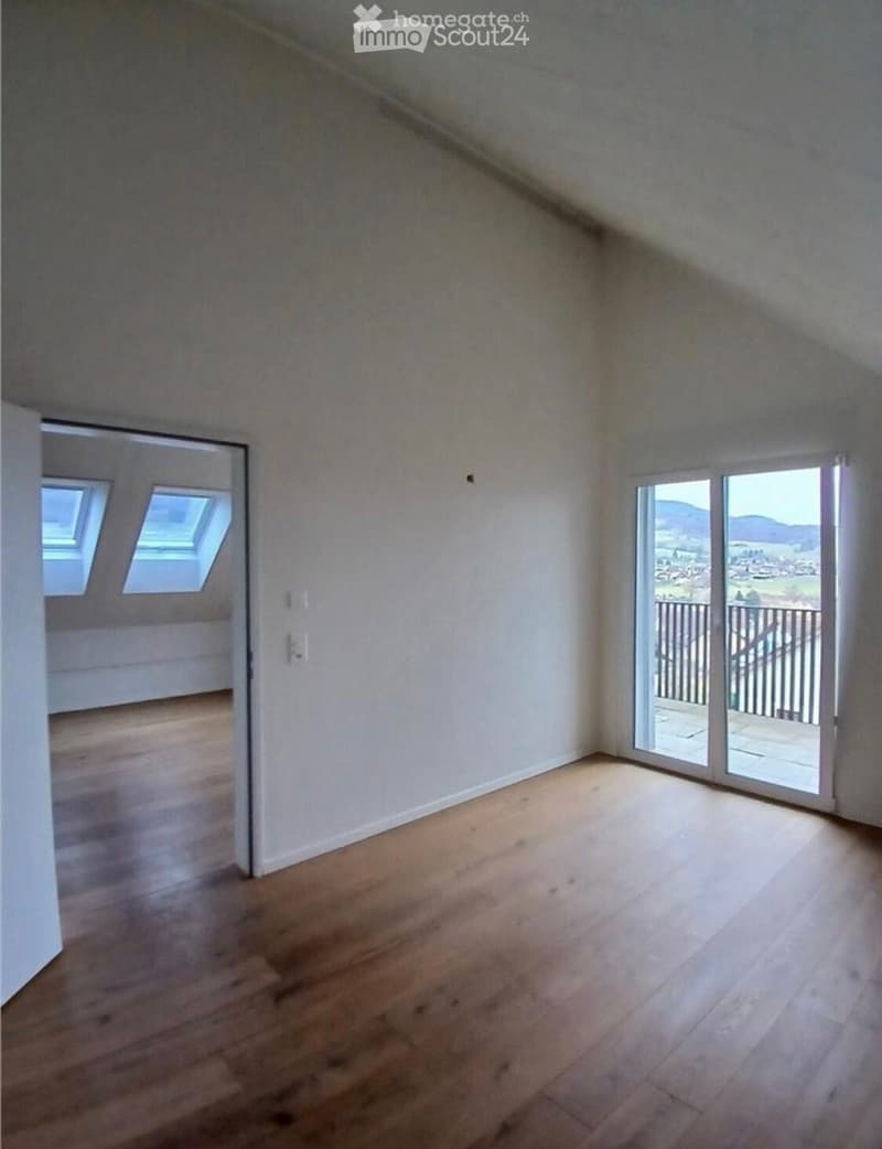 Empty room with wooden flooring, white walls, large sliding glass door leading to a balcony, and skylight windows visible in the adjacent room.