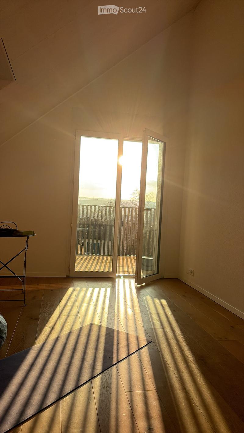 Empty room with wooden flooring and large sliding glass doors leading to a balcony. Sunlight streams in, casting shadows on the floor.