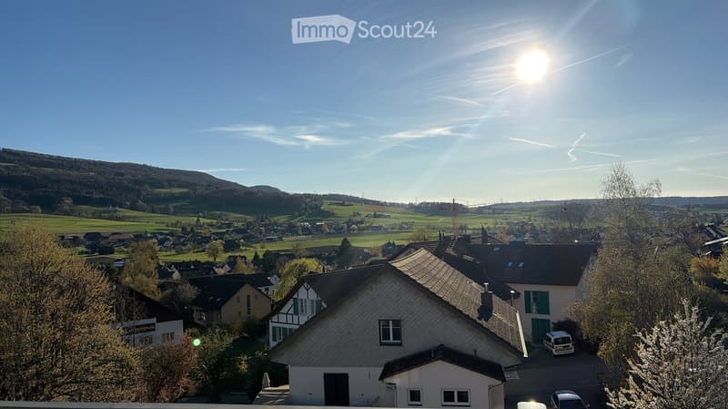 Residential neighborhood with houses, trees, and a scenic view of hills and fields under a clear blue sky.
