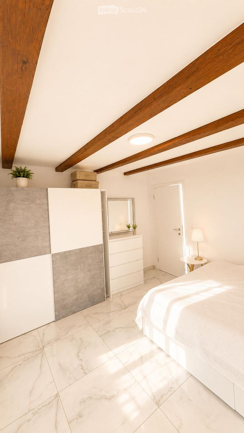 Modern bedroom with white walls, exposed wooden beams, marble flooring, a large bed, a wardrobe, a nightstand with a lamp, a small table with a lamp, and a potted plant on the windowsill.