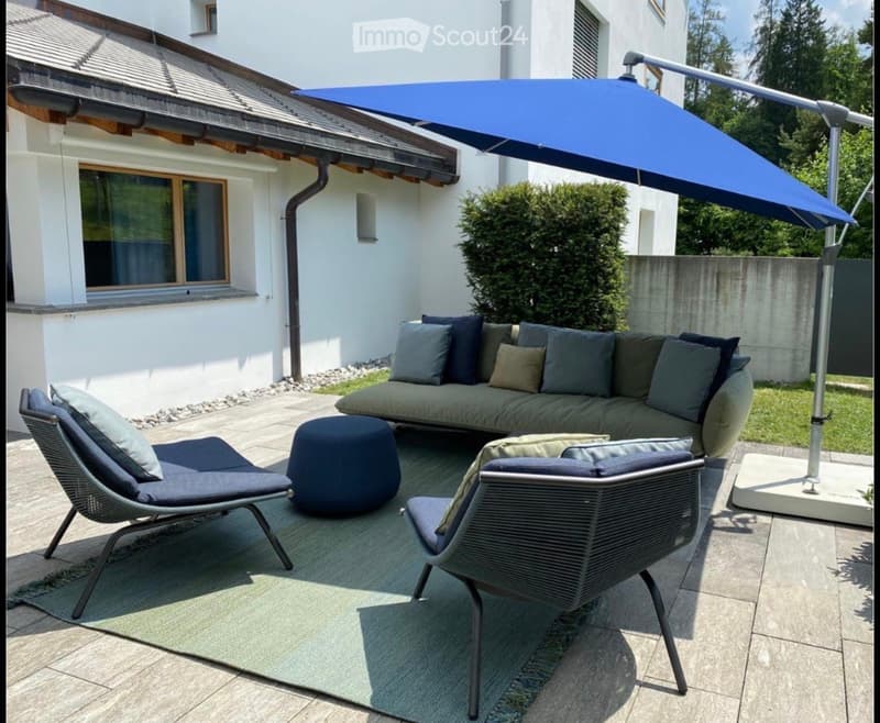 Outdoor patio with a grey sectional sofa, two wicker chairs, an ottoman, and a large blue umbrella providing shade. The area is surrounded by a white building and greenery.