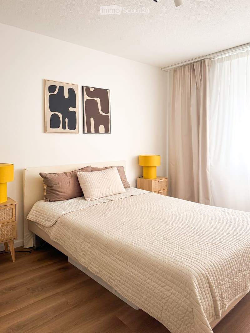 Modern bedroom with a large bed, beige bedding, two nightstands with yellow lamps, and two abstract art pieces on the wall.