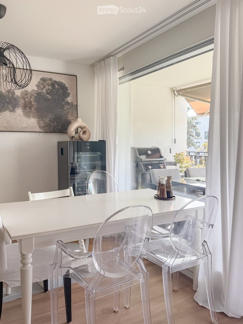 Dining area with a white table, clear chairs, a refrigerator, and a view to a balcony with a grill and outdoor seating.