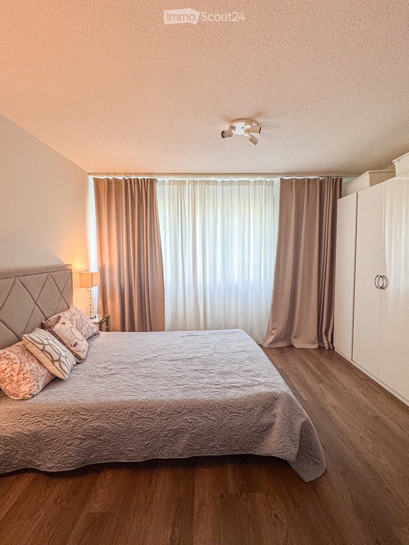 A well-lit bedroom with a large bed, decorative pillows, a nightstand with a lamp, and a wardrobe with curtains covering the windows.