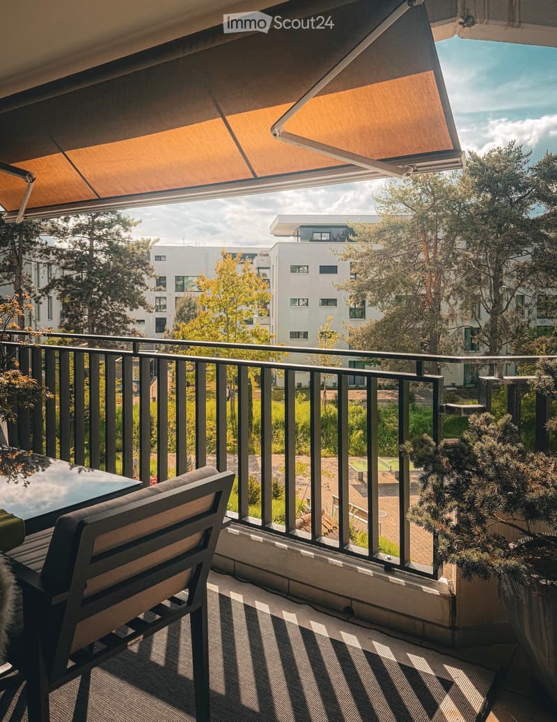 Balcony with a bench, table, and potted plants, featuring a retractable awning and a view of a modern residential area.