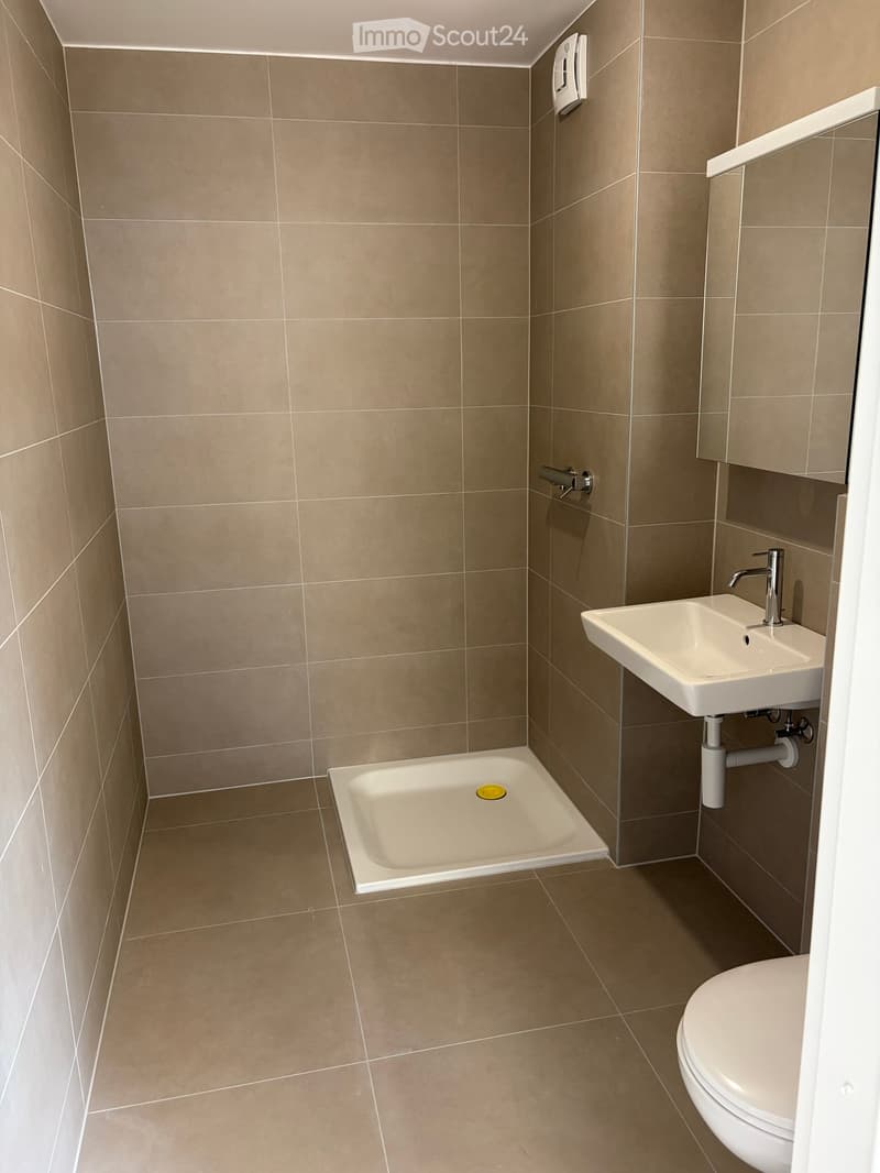 Modern bathroom with beige tiled walls and floor, featuring a toilet, sink with a modern faucet, and a small shower stall with a yellow shower cap on the floor.