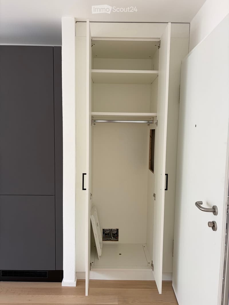 Empty built-in closet with white interior, open doors revealing shelves and a hanging rod, with a dark grey cabinet and a wooden floor visible.