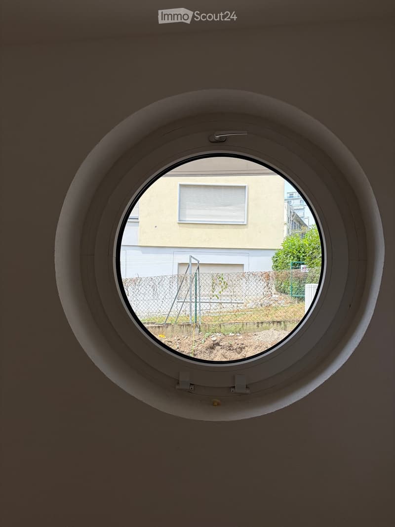 A round window with a view of a fenced yard, a building, and some greenery outside.
