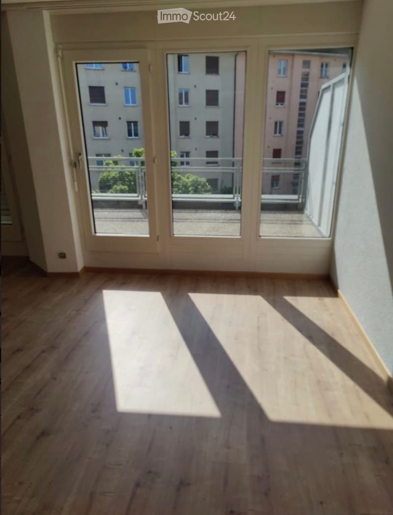 Empty room with wooden flooring and large glass doors leading to a balcony. Sunlight streams in, casting shadows on the floor.