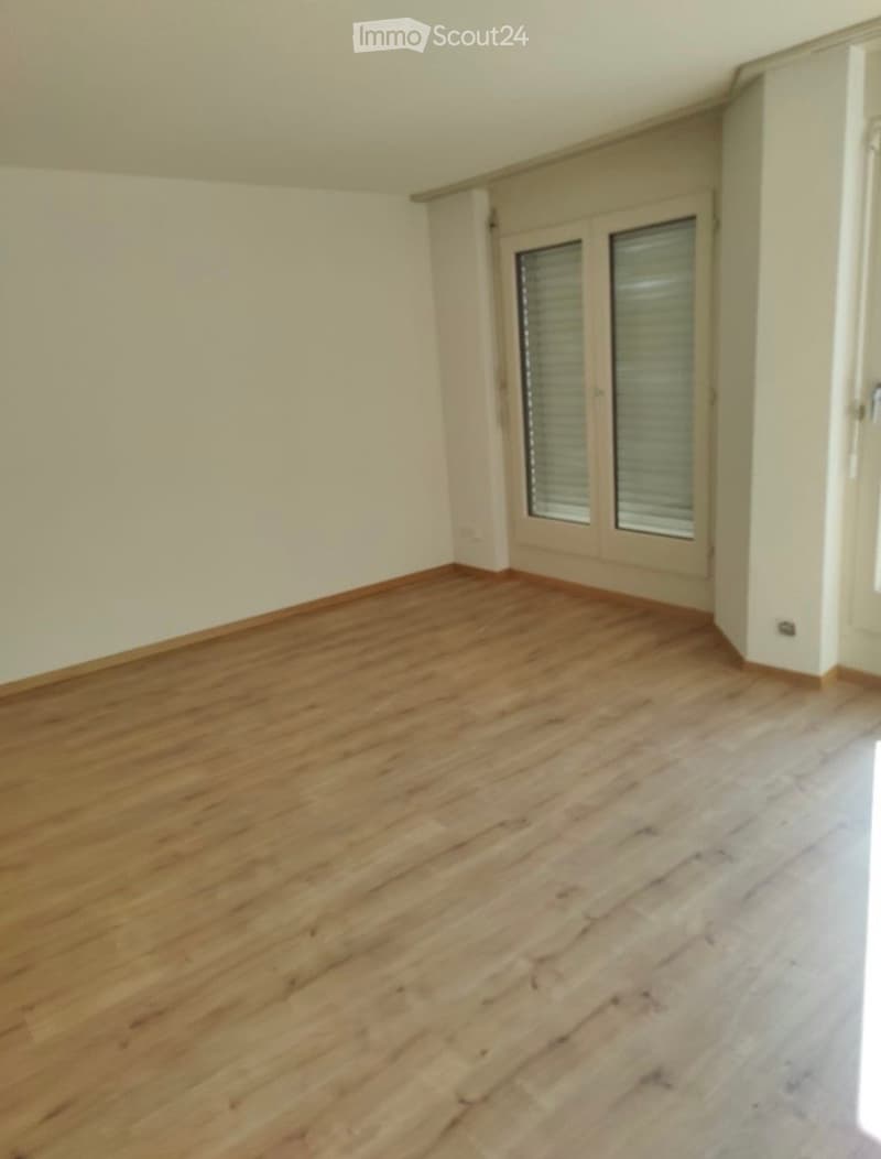 Empty room with wooden flooring and white walls, featuring a large window with shutters and a door leading to another space.