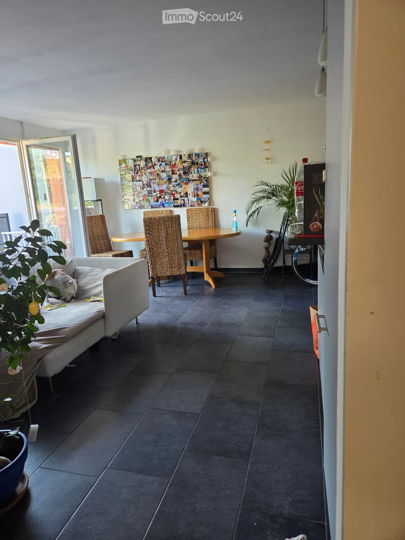 A modern living and dining area with a grey sofa, a round wooden dining table with four wicker chairs, and a large photo collage on the wall. The space features dark tiled flooring and large windows with a view of the outdoors.