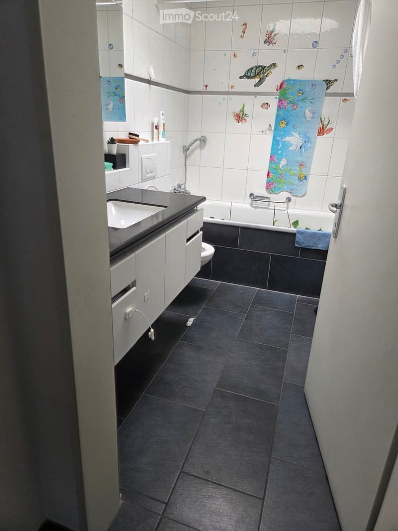 Modern bathroom with dark tiled floor, white tiled walls, a bathtub, a sink with a cabinet, and a toilet. A blue towel hangs on the side of the bathtub, and a decorative shower curtain with sea creatures is visible.