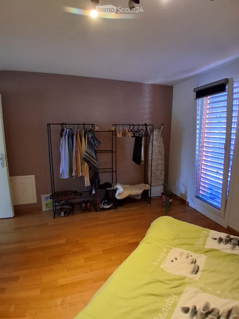 Bright bedroom with wooden flooring, a neatly made bed, a clothing rack with hanging clothes, a futon, and a small plant near the window.
