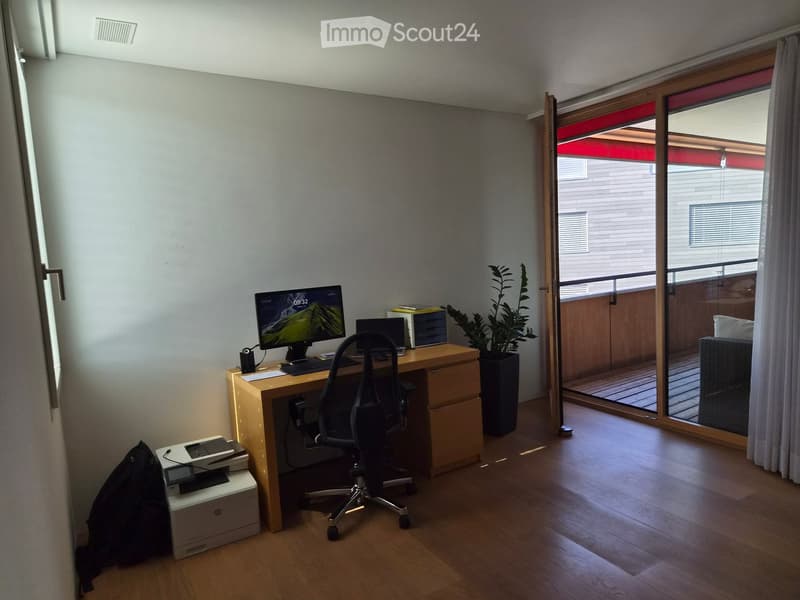 A modern, well-lit room with a wooden floor, a desk with a computer and chair, a printer, and a potted plant near a large sliding glass door leading to a balcony.