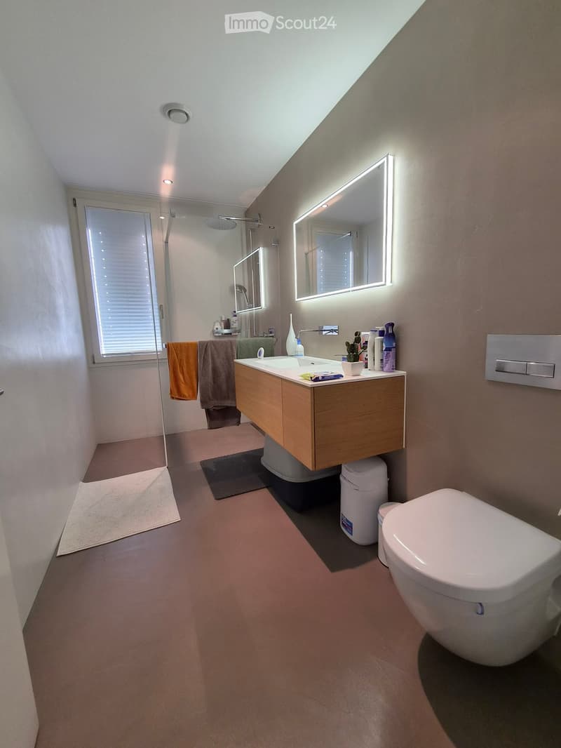 Modern bathroom with a wooden vanity, white sink, toilet, large illuminated mirror, and a glass-enclosed shower area with a window. Towels are neatly hung on a rack, and the floor is covered with a light-colored carpet.